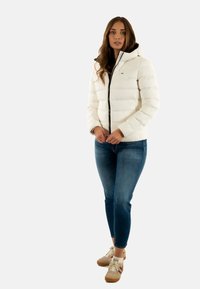 Chaqueta de plumas blanca con capucha, con un zipper negro y diseño acolchado. Combinada con vaqueros azules y zapatillas beige.