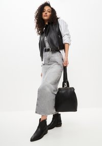 Vesta din piele neagră peste o cămașă dungată gri, asortată cu o fustă din denim gri cu talie înaltă și cizme negre până la glezne. Modelul ține o geantă neagră.