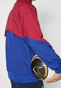 Jacka med en röd övre sektion och en blå nedre sektion, tillverkad av slät tyg. Person håller i en svart och vit fotboll med guldaccenter.