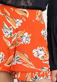 Szorty z nadrukiem kwiatowym w kolorze pomarańczowym, zdobione czarno-białymi motywami kwiatowymi. Wykonane z lekkiej tkaniny, z falbankowym dołem i dopasowanym paskiem w talii.