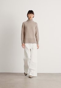 Donna che indossa un maglione beige a collo alto, pantaloni bianchi a gamba larga e scarpe beige, in piedi davanti a uno sfondo bianco uniforme.