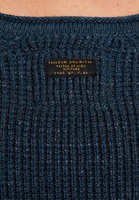 Dunkelblauer Strickpullover mit geripptem Muster und einem Etikett mit gelber Schrift, auf dem steht: "FREEDOM UNLIMITED GETESTET AUF HOHER HÖHE TYP NY C-46."
