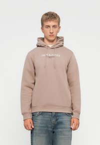 Hellbrauner Hoodie aus weichem Stoff, mit Kapuze und Zugband, mit weißem "JACK & JONES" Logo auf der Vorderseite. Getragen mit blauen Jeans.