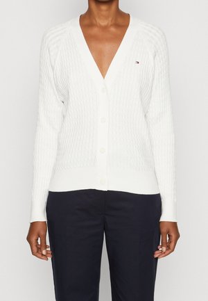 Personne portant un cardigan blanc texturé boutonné avec un petit logo sur la poitrine et un pantalon bleu marine foncé sur un fond uni.