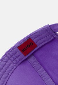 Fialová čepice s textura fabric, která má červenou štítku s nápisem "HUGO" uvnitř, umístěnou ve středu vrchu.