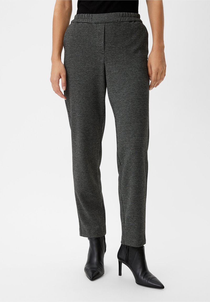 Personne portant un pantalon droit gris à carreaux, des bottines noires à talons pointus et un haut noir sur fond uni.