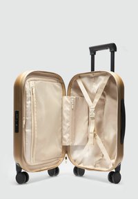 Valise rigide de couleur or métallique avec une finition texturée, intérieur beige, compartiments zippés, sangles pour sécuriser les objets et poignée rétractable.