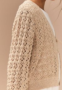 Hæklet cardigan i beige med et åbent blonderet mønster, dekoreret med sølvglimmer. Den har ribbet kant og bindebånd foran.