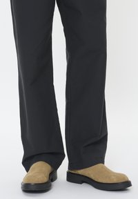 Smėliniai suede laisvi batai be raištelių su apvalia nosimi ir juodu gumininiu padu, derinami su laisvomis juodomis kelnėmis.