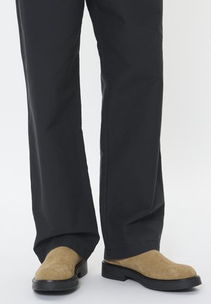 Des chaussures en daim beige sans lacets avec un bout rond et une semelle en caoutchouc noire, associées à un pantalon noir ample.