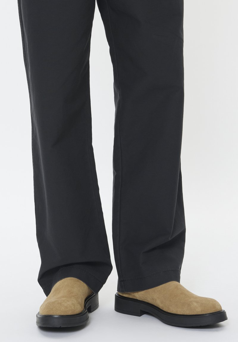 Scarpe slip-on in suede color beige con punta rotonda e suola in gomma nera, abbinate a pantaloni neri con vestibilità ampia.