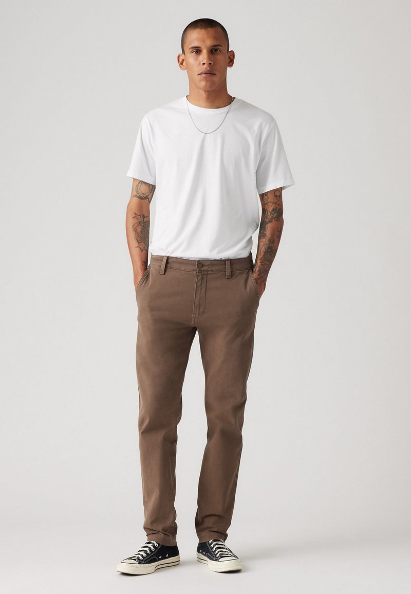 Chinos marron à coupe droite, portés avec un t-shirt blanc à col ras du cou. Des baskets noires complètent la tenue. Design simple et décontracté.