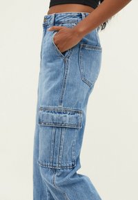 Pantalones cargo de denim azul claro con bolsillos laterales, de cintura alta y corte holgado. La tela tiene una textura con detalles de costura visibles.