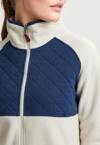 Vilnas jaka ar krēmkrāsas ķermeni un tumši zilu rūtainu krūšu paneli, augstu apkakli un rāvējslēdzēju ar brūnu ādas cilpu.