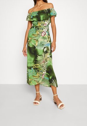 Kobieta ubrana w zieloną sukienkę midi z odkrytymi ramionami, ozdobioną tropikalnym wzorem liści i kwiatów, zestawioną z białymi sandałami na obcasie na białym tle.