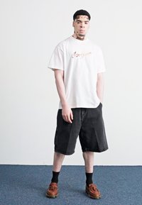White t-shirt with “Carhartt” logo, black denim shorts, black socks, brown leather shoes. Standing on a blue carpet against a plain background.