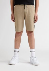 Beige shorts med en glat tekstur, der har en sidelomme og et logopatch, kombineret med hvide sneakers og stribede crew-sokker.