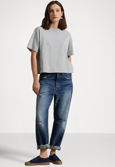 Žena v nosenej voľnej sivej krátkosleevej blúzke, roztrhaných modrých džínsach s manžetami a námorníckych modrých teniskách stojaca pred jednoduchým pozadím.