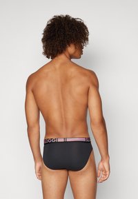 Schwarze Unterhose im Brief-Stil mit einem Sloggi-Logo-Band in Pink und Lila. Glattes, dehnbares Material mit einer enganliegenden Passform, von hinten betrachtet.