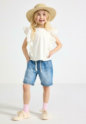 Ung pige smilende, iført en stråhat, hvid bluse med flæser, blå denimshorts, lyserøde sokker og beige sneakers, stående med hænderne på hofterne.