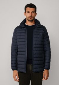 Mężczyzna ubrany w ciemnoniebieską pikowaną kurtkę z podniesionym kołnierzem, czarny sweter i ciemne spodnie, stojący na tle jasnej, jednolitej ściany.