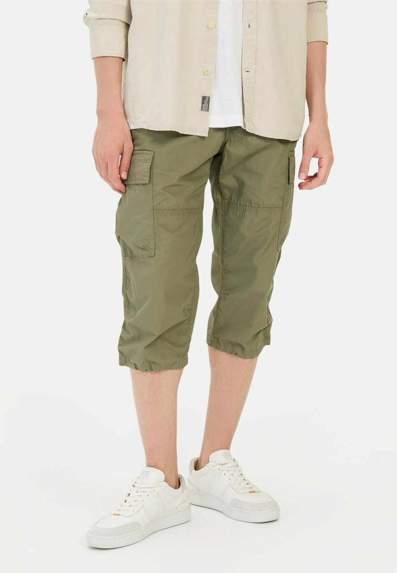 Olivegrüne Cargo-Shorts mit einer entspannten Passform, knielang und mit elastischen Bündchen. Verfügt über mehrere seitliche Taschen. Kombiniert mit weißen Sneakers.