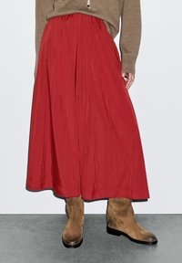 Jupe maxi plissée rouge en tissu léger, associée à des bottes chelsea en daim tan ; ourlet simple et texture lisse.