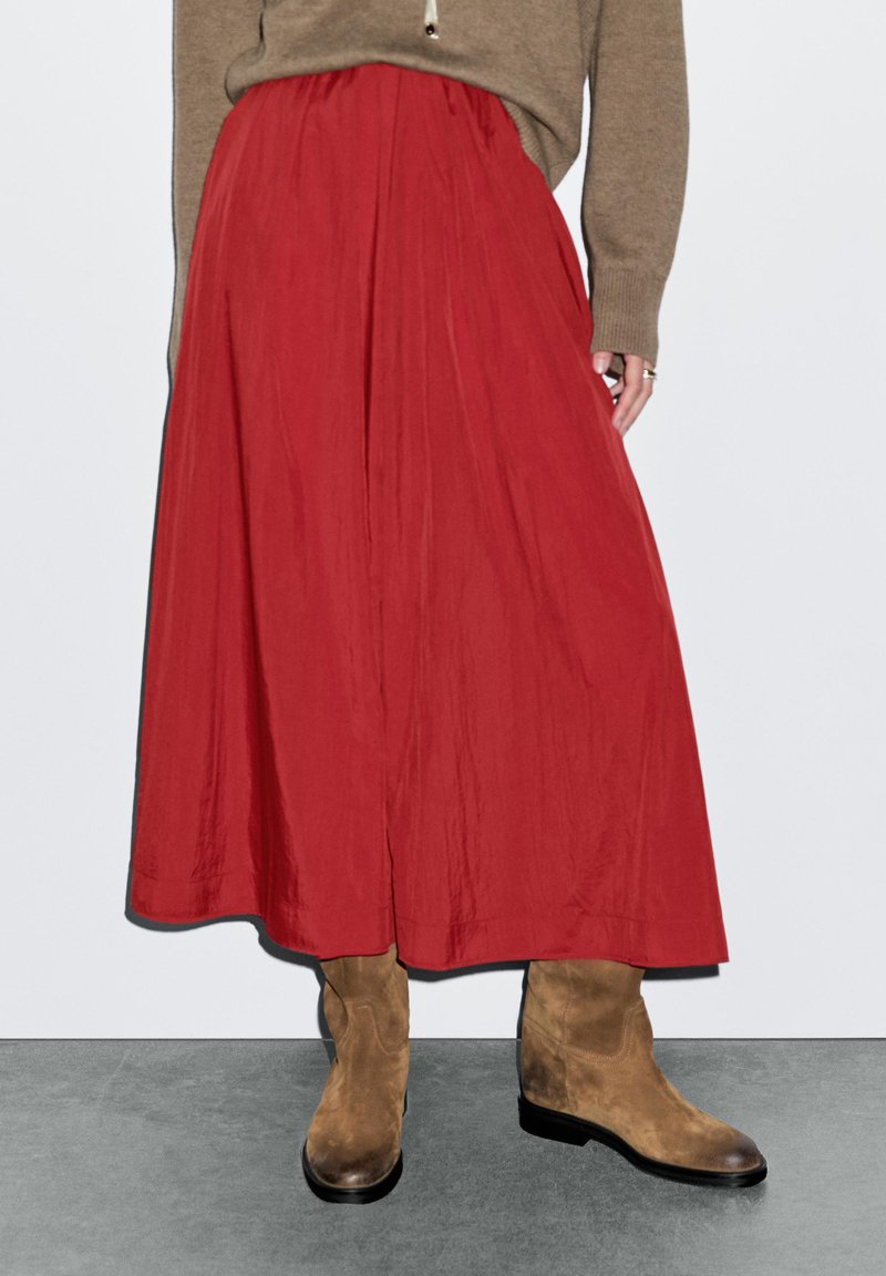 Jupe maxi plissée rouge en tissu léger, associée à des bottes chelsea en daim tan ; ourlet simple et texture lisse.