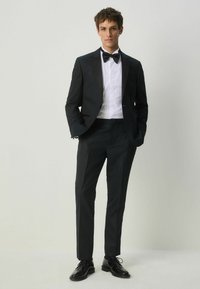 Fekete szmoking texturált anyagból, selyem hajtókával, fehér dressz inggel és csokornyakkendővel. Slim-fit nadrág, fényes fekete dressz cipővel párosítva.