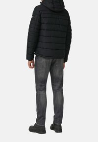Zwarte pufferjack met horizontaal gestikte patroon, capuchon, gecombineerd met grijze denim jeans en donkere laarzen, gezien van achteren.