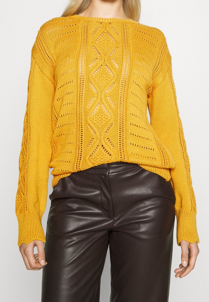 Gul stickad tröja med ett texturerat mönster, med kabeldesign och öppna maskor, i kombination med mörkbruna åtsittande byxor.