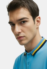 Polo shirt azzurro chiaro con vestibilità slim, con colletto a righe blu navy e giallo e apertura con due bottoni. Texture liscia.