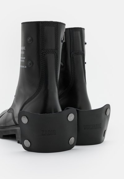 Black leather ankle boots with a smooth finish, featuring prominent zippers, reinforced stitching, and rubber heel caps embossed with "ZADIG" and "VOLTAIRE."