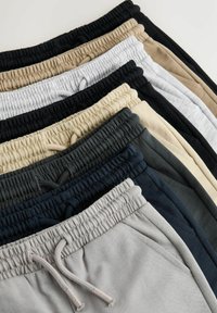 Pile de pantalons de survêtement à taille élastique en différentes couleurs neutres, montrant la partie supérieure avec cordons de serrage et poches latérales.