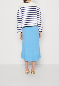 Kvinna som bär en vit kort tröja med blå ränder, en ljusblå midikjol och beige högklackade sandaler, stående med ryggen vänd framåt.