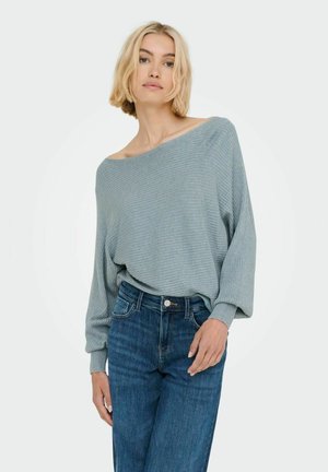 Suéter ribeteado en gris claro con diseño de hombro caído, corte oversized y mangas largas, combinado con jeans azul oscuro.