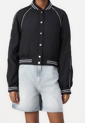 Sort bomberjakke med hvide piping og stribede ærmekanter kombineret med lyseblå højtaljede denimshorts på en stående person.