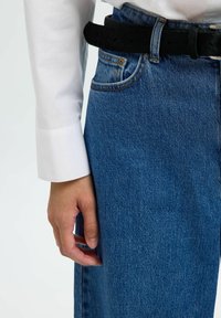 Vaqueros de mezclilla azul con un corte holgado, cintura alta y un cinturón negro. Los puños de una camisa blanca son visibles, contrastando con la textura de los vaqueros.