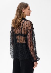 Czarna koronkowa bluzka z długimi rękawami, zdobiona skomplikowanymi wzorami kwiatowymi i przezroczystym tyłem. Teksturowany materiał dodaje głębi i szczegółowości.