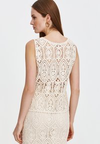Kremowa koronki bluzka z misternymi wzorami kwiatowymi i liściastymi, bez rękawów, z falbaniastym dekoltem, prezentująca teksturowane wykończenie.