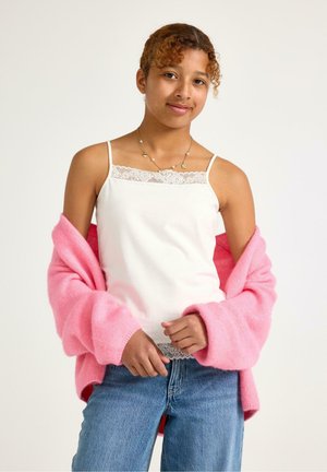 Giovane donna che indossa una canottiera bianca con bordatura in pizzo, jeans blu e un cardigan rosa larghetto sulle spalle, sorridendo dolcemente.