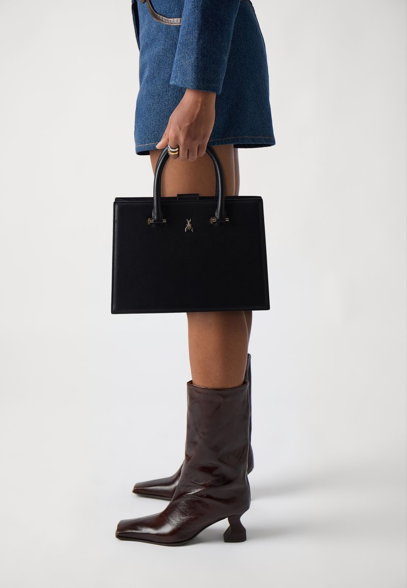 Black leather handbag with structured design, short handles, and gold hardware; paired with knee-high brown leather boots.