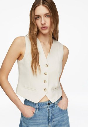 Junge Frau mit langen hellbraunen Haaren, trägt eine ärmellose cremefarbene Weste mit Knöpfen und blaue Jeans, Hände in den Taschen, neutraler Hintergrund.