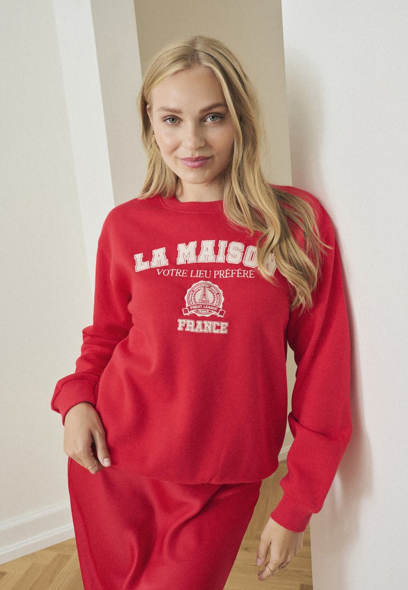 Felpa rossa con testo bianco che recita "LA MAISON VOTRE LIEU PRÉFÉRÉ FRANCE", con vestibilità comoda e maniche lunghe. Texture liscia.