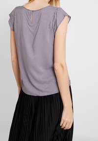 Kortärmad topp i ljuslila med geometriskt mönster, med rund hals och en liten knappdetalj på ryggen. Tyget ser slätt och lätt ut.