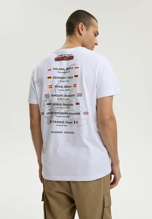 Mężczyzna w białej koszulce z kalendarzem mistrzostw Super Enduro 2025/2026 oraz nazwami europejskich miast, datami i flagami na plecach.