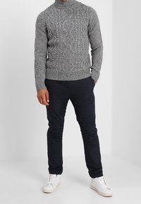 Pull à col roulé gris en maille torsadée associé à un pantalon slim bleu marine foncé. Le mannequin porte des baskets blanches aux semelles beiges. Design simple.