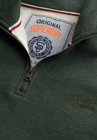 Sudadera verde con cremallera, que presenta un tejido texturizado, un cuello en contraste con ribete rojo y un logotipo bordado en el pecho y la etiqueta.