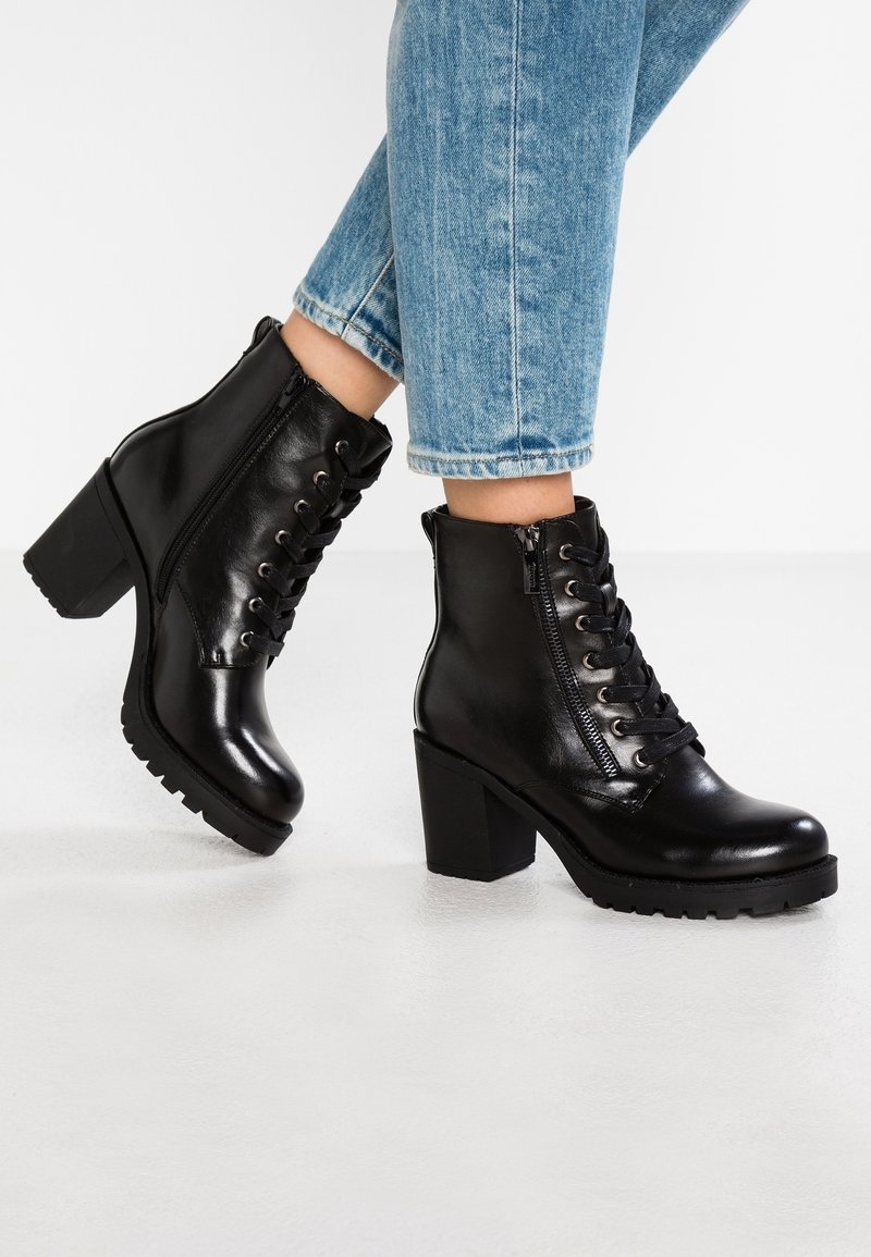 Bottines noires à talons hauts avec lacets et fermeture éclair latérale, portées avec un jean bleu clair retroussé, posées sur une surface blanche.