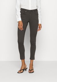 Jeans ajustados de color gris carbón con un dobladillo recortado, cinco bolsillos y cierre de botón, combinados con una camisa blanca de botones y chanclas negras.